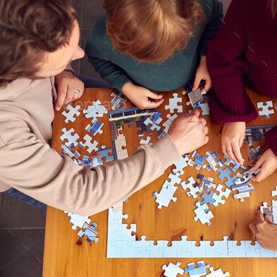 Overhead Shot Of Family Sitting Around Table At Home Doing Jigsaw Puzzle Together